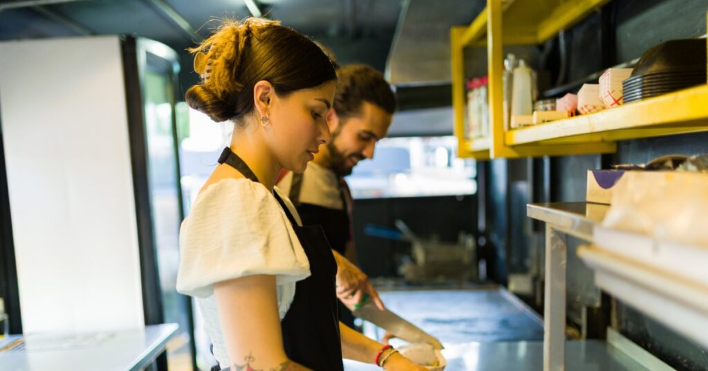 Two people preparing food in a compact kitchen with yellow shelves, containers, and bright window light nearby.