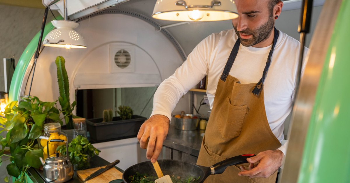 A person cooking at a counter inside a food truck with plants, utensils, green walls, and overhead lights.