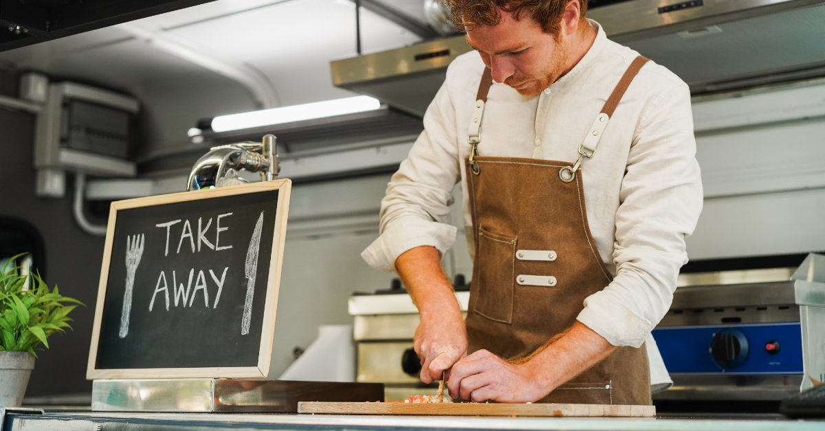 A person preparing food on a counter near a chalkboard take-away sign inside a compact kitchen with overhead lights.
