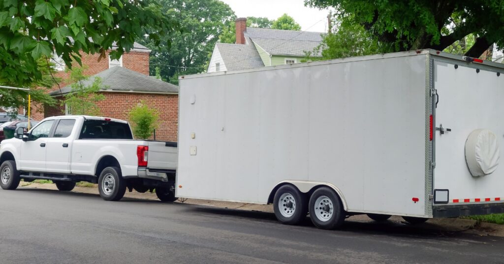 A large white utility trailer connected to a white pickup truck parked on the side of a residential road.