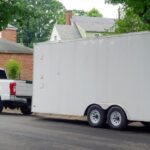 A large white utility trailer connected to a white pickup truck parked on the side of a residential road.