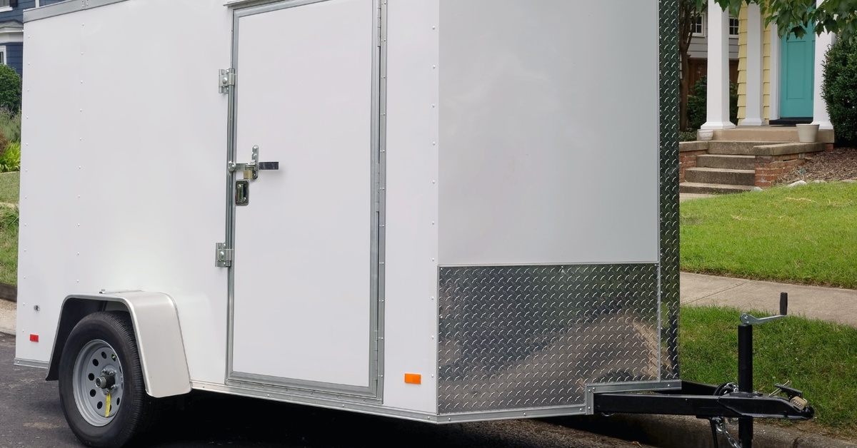 A close-up of a single-axle white utility trailer with a side door parked on the side of a residential road.