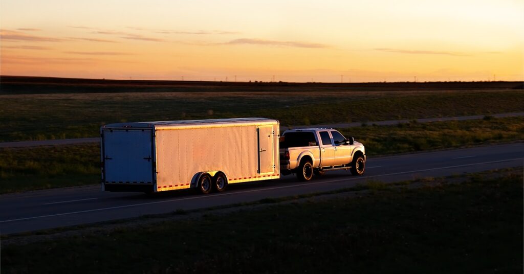 A pickup truck hauling a large enclosed trailer along an empty highway surrounded by fields at sunset.