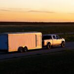 A pickup truck hauling a large enclosed trailer along an empty highway surrounded by fields at sunset.
